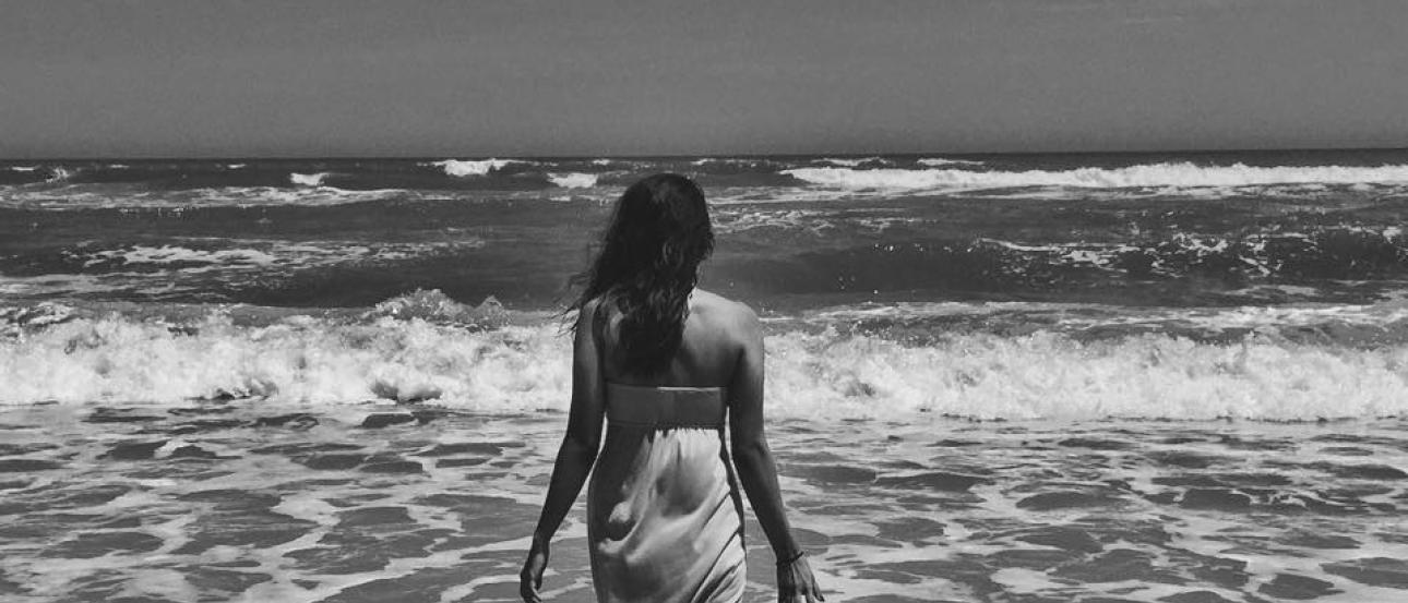 A woman wades in the ocean, wearing a dress, seen in black and white. This alt text was added with Al; accuracy may vary.