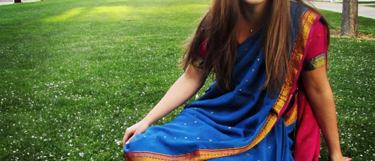 Smiling woman in blue and red attire sitting on green grass near buildings. This alt text was added with Al; accuracy may vary.