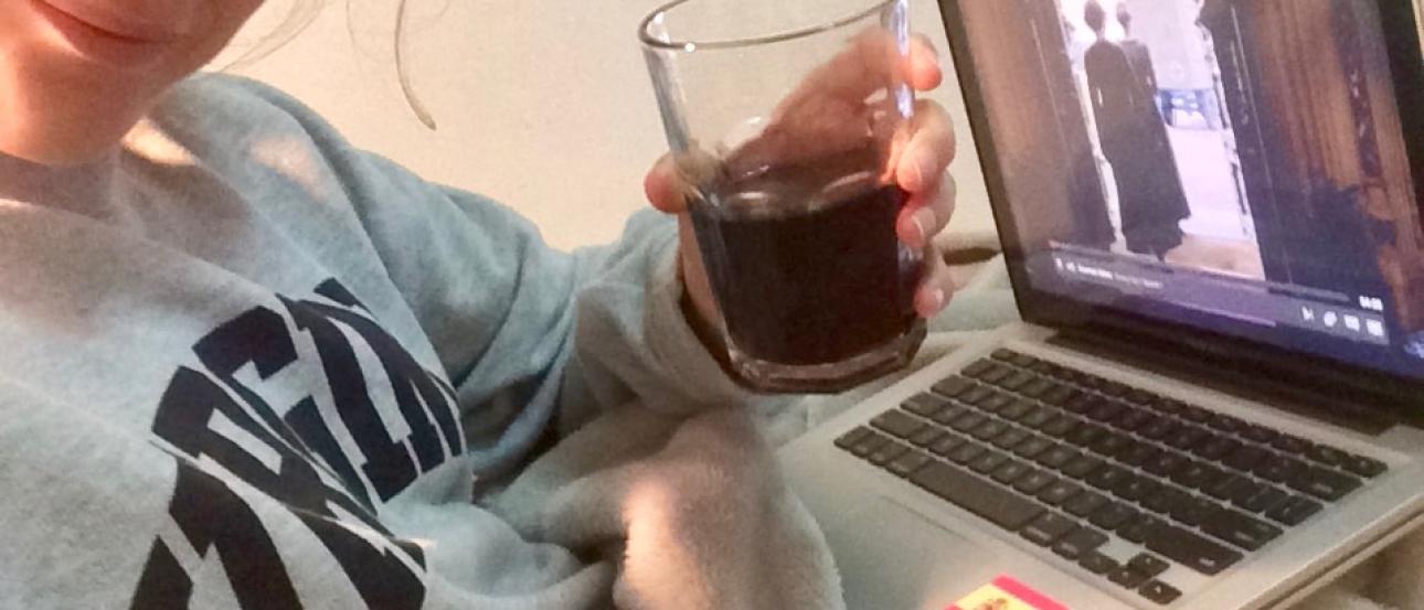 Person smiling, holding a drink, watching a show on a laptop. Cozy setting. This alt text was added with Al; accuracy may vary.