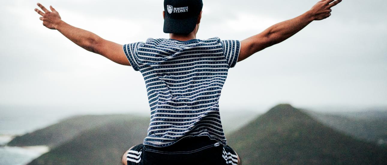 Sitting person with arms outstretched on a railing, overlooking coastal hills. This alt text was added with Al; accuracy may vary.