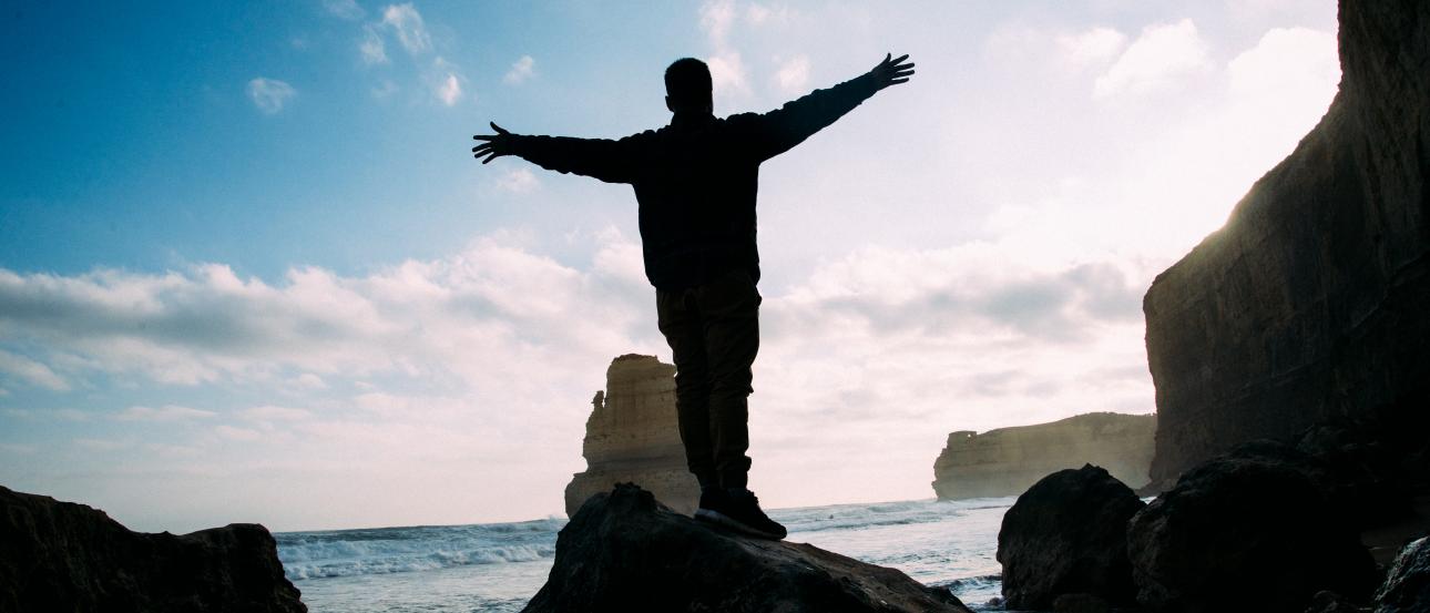 Silhouette of person on rocks by the sea, arms outstretched, cliffs in the background. This alt text was added with Al; accuracy may vary.