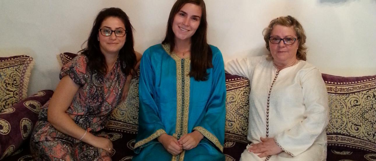 Three women in festive attire sitting on a patterned couch, smiling indoors. This alt text was added with Al; accuracy may vary.