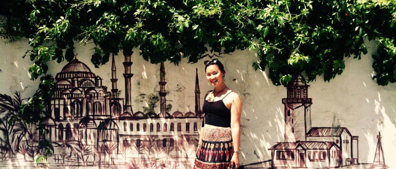 Woman stands by mural of Istanbul skyline, greenery overhead, sunny day. This alt text was added with Al; accuracy may vary.