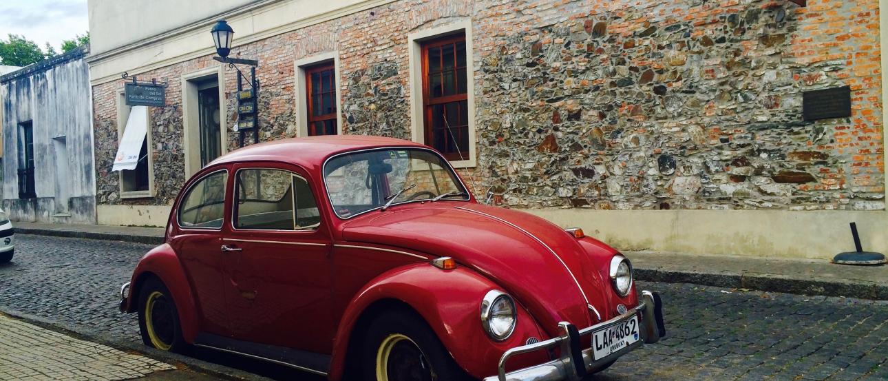 Red vintage Volkswagen Beetle parked on cobblestone street by a brick building. This alt text was added with Al; accuracy may vary.