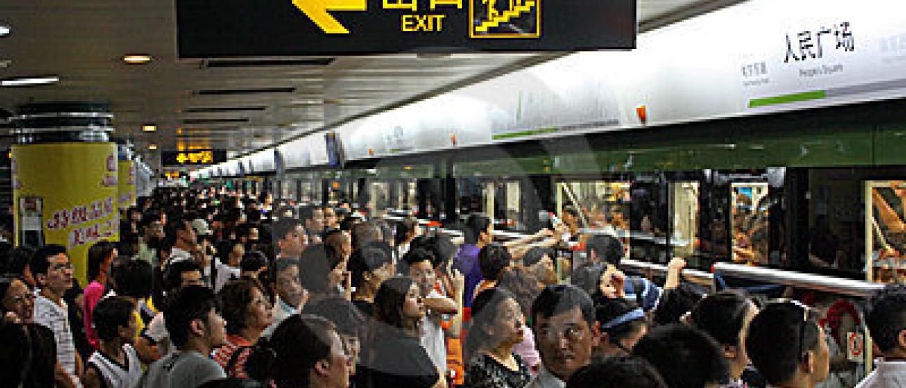 Crowded subway platform with people waiting for a train, exit sign visible above. This alt text was added with Al; accuracy may vary.