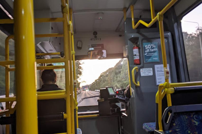 Image shows perspective of sitting on a bus seat looking down the aisle towards the front of the bus, where sunrise is visible through the windshield.