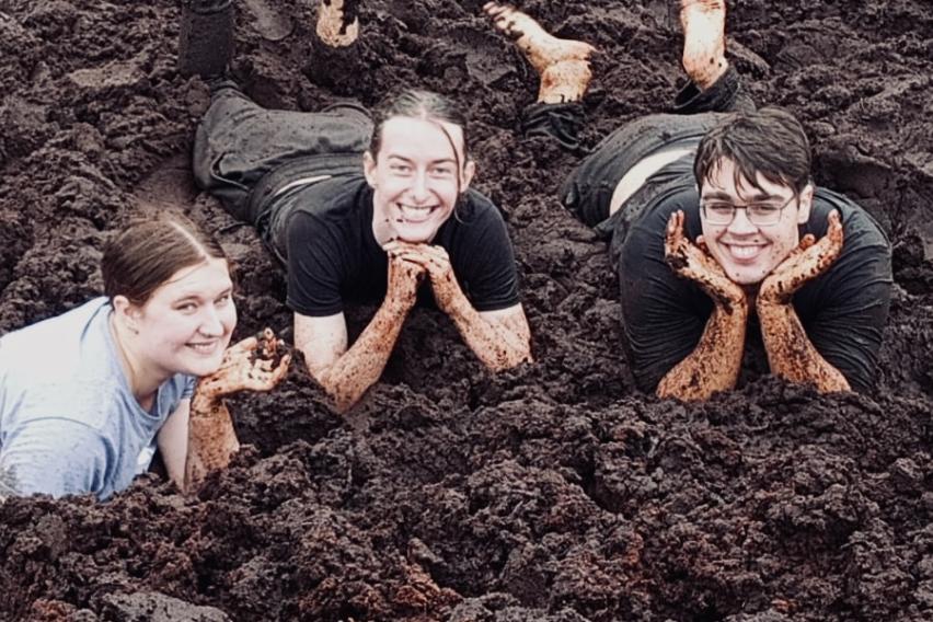 3 students lie in a bog facing the camera. They are smiling, with their hands under their chin. They are covered in mud.