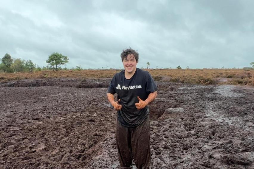 Short white man, dressed in dark muddy clothes, standing by a bog. He is smiling with two thumbs up