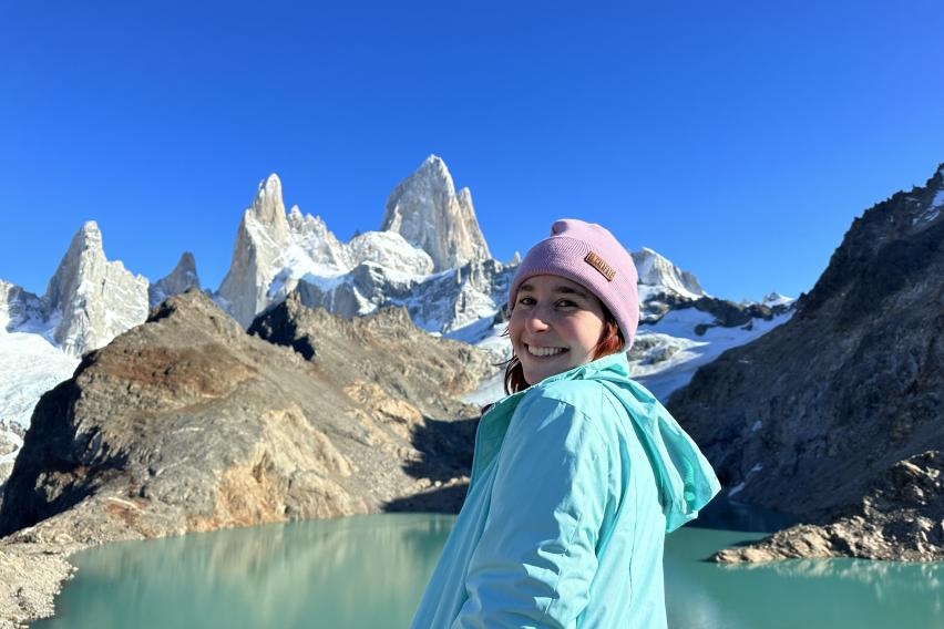 student wearing jacket and knit hat smiling in front of a body of water that backs up to a snow capped mountain