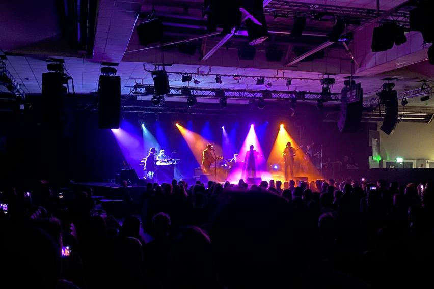 Gregory Alan Isakov, second from left, alongside his bandmates onstage, each in a different-colored spotlight. From the left is piano (blue), Gregory on guitar (orange), violin (pink), and bass guitar (yellow/orange). There is a sea of heads in front of them, an audience.