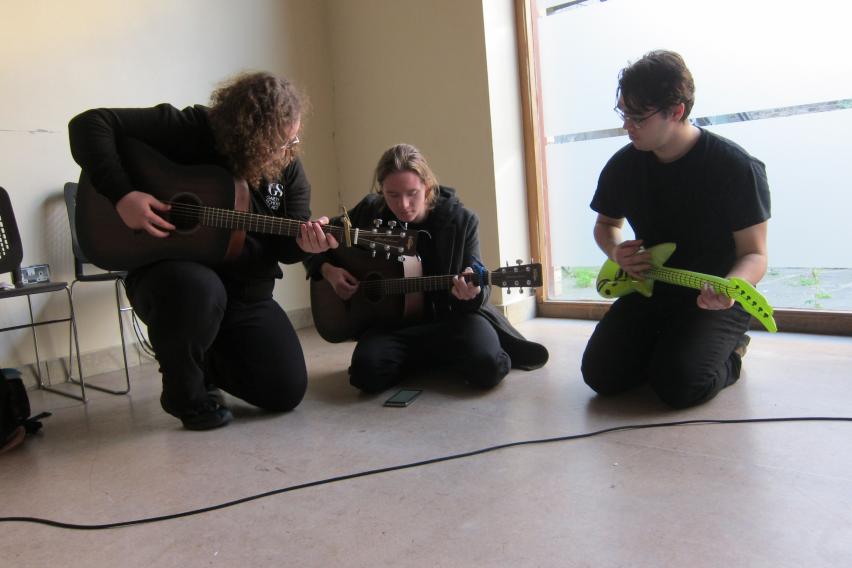 Three students sit with guitars in hand, looking at a phone screen. The student on the far right is holding a green inflatable guitar.