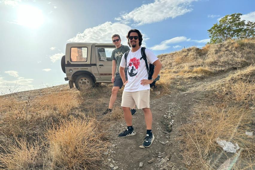 two students standing on dry grassy terrain walking down a slight decline away from the sun and a jeep