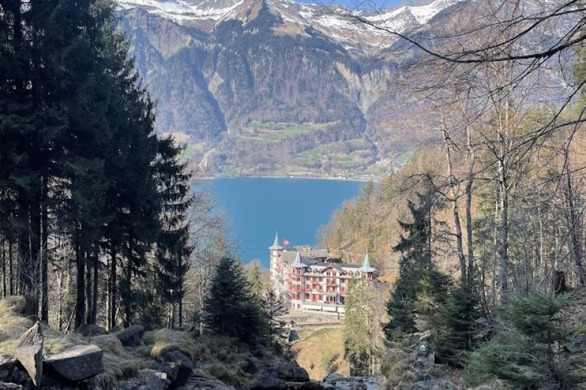 View of the lake in Brienz, an old hotel, and a massive mountain against a blue sky. Picture taken from a high elevation.