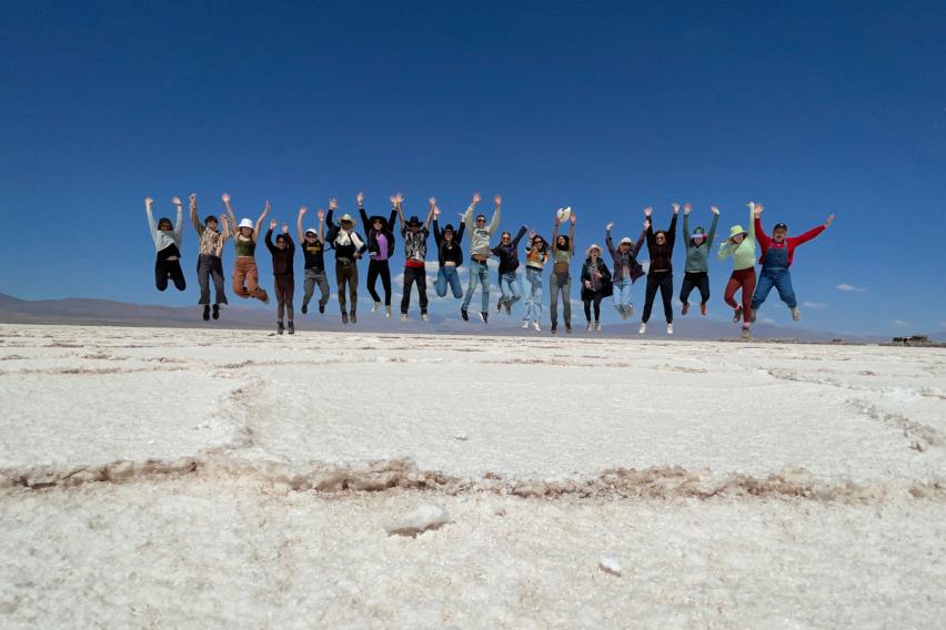 A large group of people jumping in unison with arms raised on a vast white salt flat under a clear deep blue sky