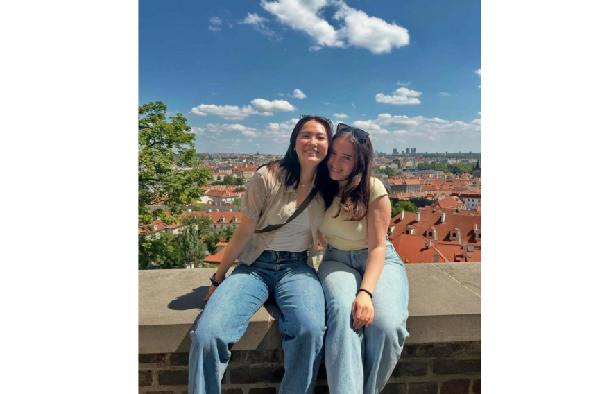 2 female students sitting on a stone wall, on a sunny day and beautiful city backdrop