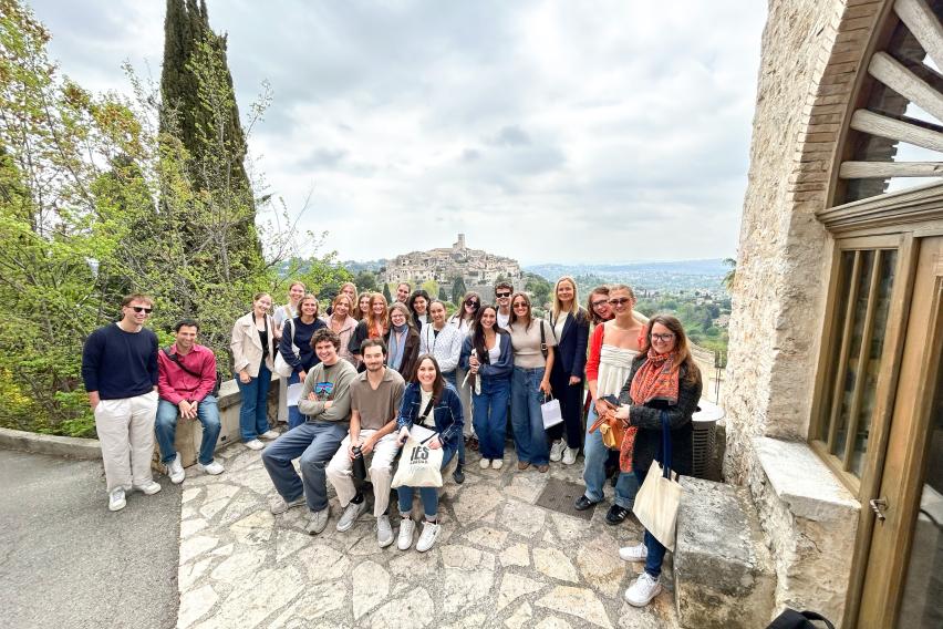 A group of IES Abroad students stands on a stone terrace beside a historic building, with trees and a hilltop town in the distance under a cloudy sky.