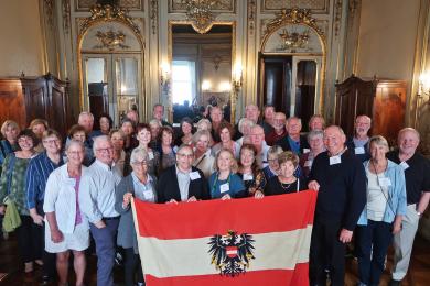 A group of older individuals standing in front of a mirror with the flag of Vienna and smiling for the camera.
