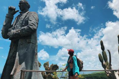 A student looks at a bronze statue of Charles Darwin, about twice her height, in the Galápagos Islands, Ecuador. In the background, there’s a body of water and cacti.
