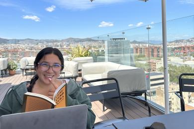 student holding book in front of laptop at table on rooftop with umbrella and cityscape in background