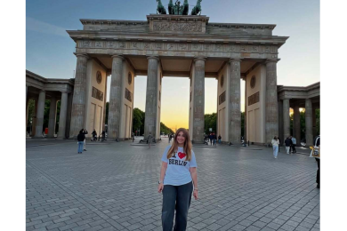 A girl standing in front of the Brandenburg Gate in Berlin, wearing a t-shirt that reads "I love Berlin", with the sunset lighting up the sky.