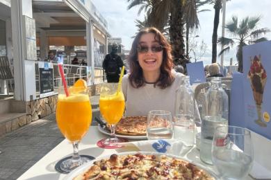 Girl in sunglasses smiling at camera sitting at table outside with orange juice and pizza