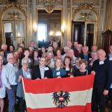 A group of older individuals standing in front of a mirror with the flag of Vienna and smiling for the camera.