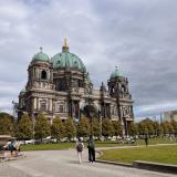 Two tourists stop to take photos in front of the Berlin Cathedral, a Baroque-style palace with a green center dome and four corner towers.