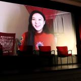 A video of Hyunseo Kong giving her acceptance speech plays on a large theater screen, while the IES Center Director holds the trophy.