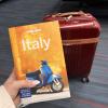 Image of a hand holding up an orange guidebook titled Italy with a maroon suitcase in the background.