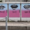 Three electrical boxes that have been spray painted, fading from light blue to purple, on each box there are lips in different positions mouthing out the word VAG 