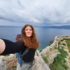 Ginger lady smiling into a camera at the top of a hill in front of the Monemvasia sea.