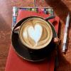 Artful cappaccino with heart-shaped foam, red notebook, pencil, sunny café table. 