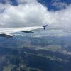 The right wing of a plane cuts across the sky, as viewed from the window seat. Cloudy blue skies look down on a shadowy mountain terrain.