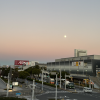 Picture of a sunset by a train station and road, the moon is full and the sky is a gradient of pale orange to blue
