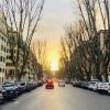 A view down a Roman street at dusk. There are cars parked on the side and the sun sets in the middle.