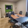 A small meeting room with a wooden conference table, office chairs, a wall-mounted TV, and a green moss accent wall