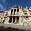 A large, tan building with pillars and detailed architecture with a Chilean flag stands against a blue sky filled with clouds. 