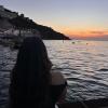 A person sits by the water in Sorrento, watching the sunset over the sea as the sky is pink and orange. 