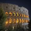  Illuminated view of the Colosseum in Rome at night, with its arches glowing yellow against the dark sky and trees in the foreground.