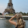 A girl standing in front of the Eiffel Tower with her hands over her eyes as if she is looking through binoculars.