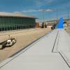 Airplane wing with a bright blue wingtip on an airport tarmac. The photo is taken from a window seat near the wing, showing the long white wing stretching toward the distance, with flap panels visible along its trailing edge. Below the wing is a concrete apron with a red guideline and some darker stained areas. Near the center-bottom of the image there is a small white and yellow ground vehicle positioned close to the wing, with a tow bar extended toward the aircraft. To the left side of the scene is an air