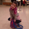 Young girl smiling while sitting on a small purple ride-on suitcase in an airport terminal.