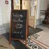 A sign on a clapboard reads "Welcome! We're open. Come on in" and has a cartoon coffee cup drawing. An open door is behind it.