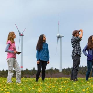 4 people in a field of flowers looking into the distance, wind turbines are behind them