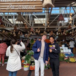 two students holding a thumbs up in front of a food market with signs 