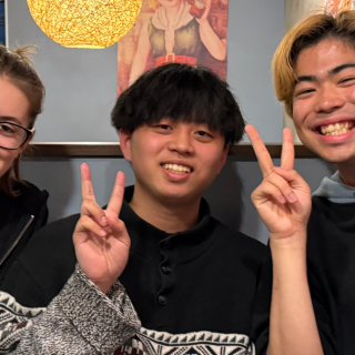 Three friends smiling and holding up peace signs while eating in a restaurant booth in Tokyo, Japan