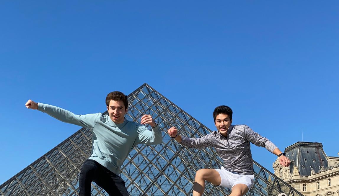 two students jump into a pose for a photo in front of the Louvre