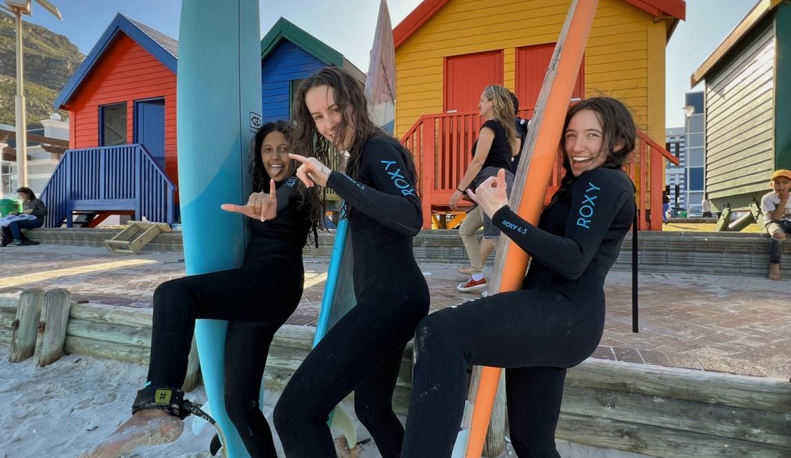 three students pose for a fun photo on the beach with surf boards in Cape Town
