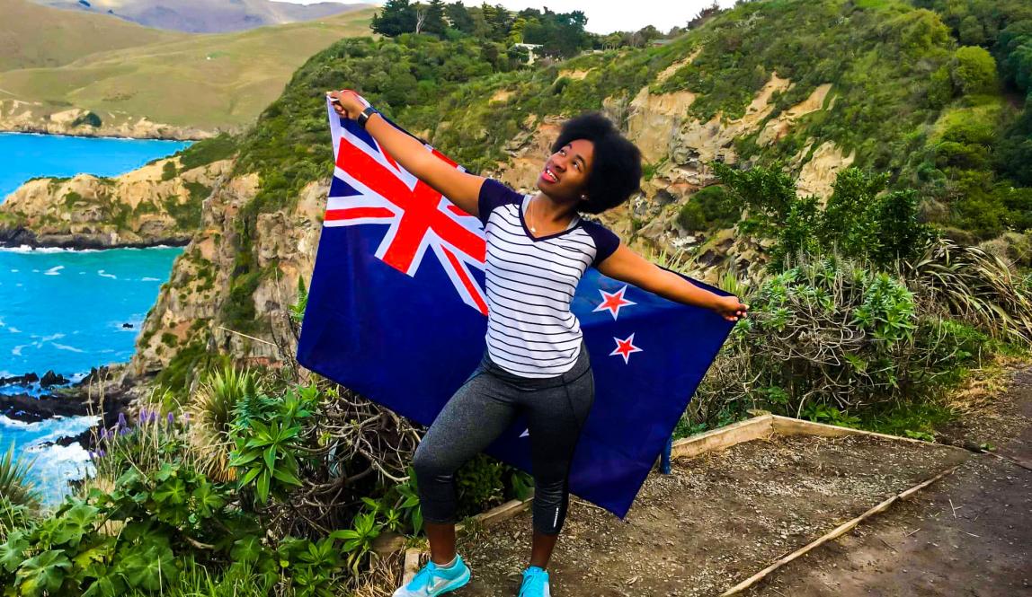 a student stands on the New Zealand coast posing with a New Zealand flag