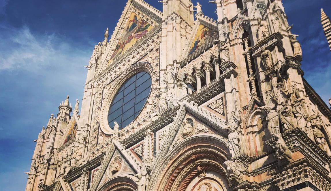 A cathedral in Siena, the Duomo Di Siena, on a sunny day. The exterior is an off-white color, with ornate details on the structure.